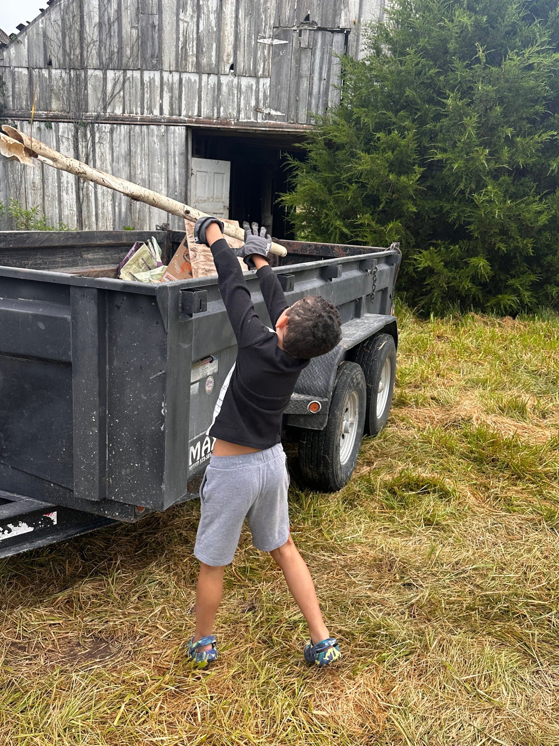 A child lifting a pole into the dump trailer beside a weathered barn